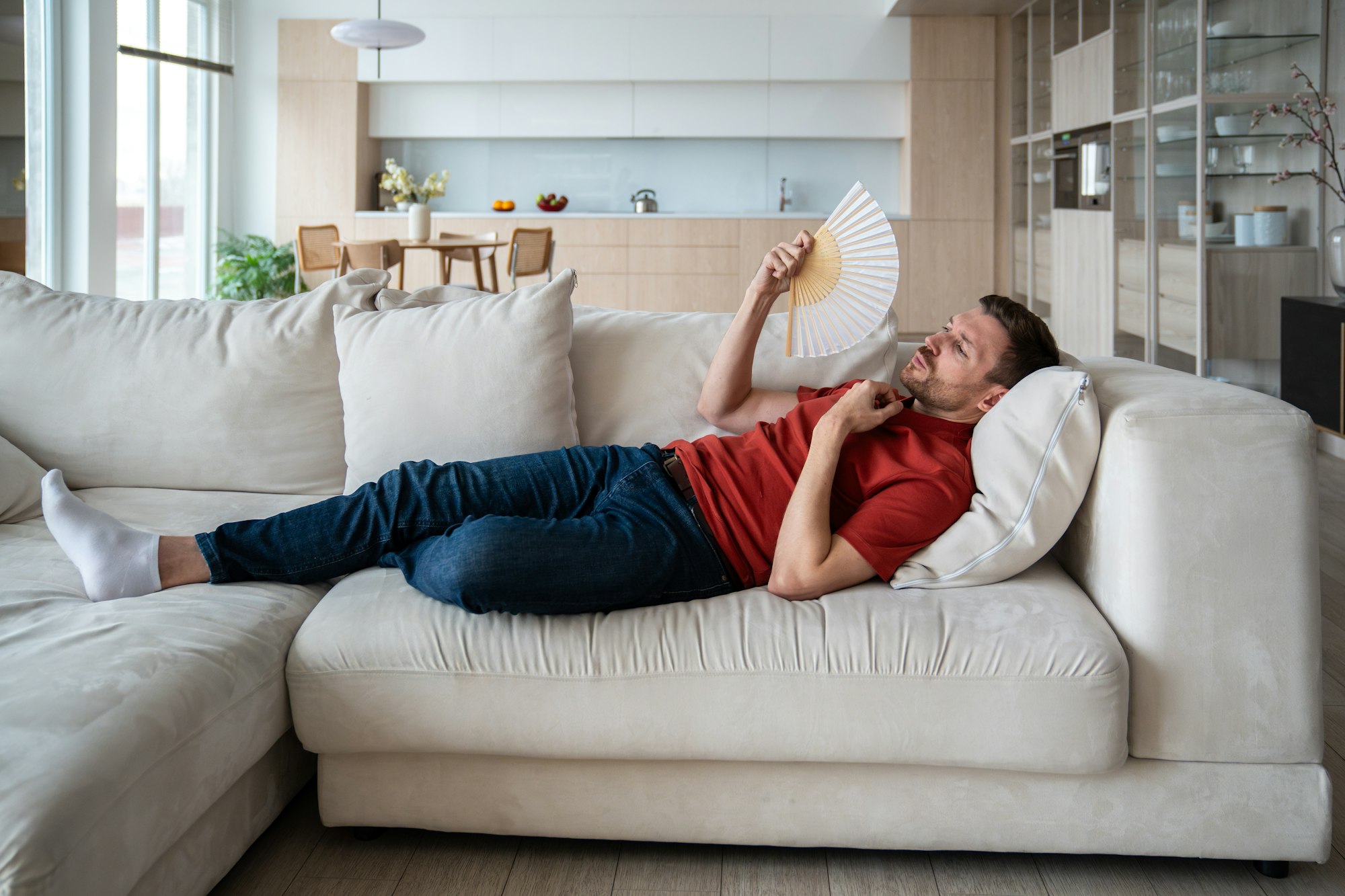 Exhausted man with difficult breath lying on sofa, waving with hand fan in need of cool, fresh air